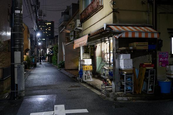 A narrow alley at night featuring a small shop with a striped awning. The scene includes wet pavement reflecting light, surrounding buildings, and people in the distance. Street lights and power lines are visible.