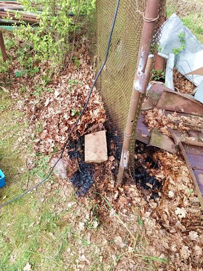 Rusty fence post with newly tacked on gate hinges made from more rusty scrap metal. There are dried leaves all along the fence. The pile on the bottom of the fence post looks suspiciously black and scorched. A bunch of metal trash is mixed with more leaves on the side, which needed removing and wetting in case of embers.