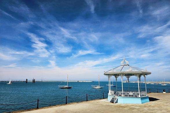 A white and light blue bandstand stands on a stone pier overlooking a calm, blue sea dotted with sailboats under a partly cloudy sky.