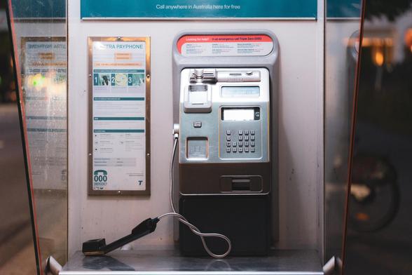 The image shows a public payphone. It includes a handset, dial pad, and display screen, with instructions visible on the side. It's installed in a booth, and there's a sign indicating it's for making calls, possibly in Australia given the wording at the top.