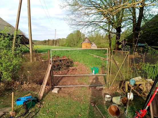 New (recycled) meadow gate fitting on its post, somewhat crooked but much better than the old, which is still leaning on the fence next to it. There is, almost invisible in this foto, a green wire fence installed in the pipe frame now. If you look even closer, you can see the fence had lots of breaks which have been carefully mended with old wire. On the ground rests a blue tool tote, a green welder, Mjölnir the post pounder and some old pots and buckets that were pulled from the trash pile. The steel trash pile has moved further out and where it was is now a freshly raked and wet patch of ground. The burnt leaves have gone to a less flammable neighbourhood.