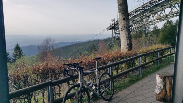 Rennrad steht an der Bergstation an ein Geländer gelehnt. Im Hintergrund im Tal ist andeutungsweise Freiburg zu erkennen. Davor Apparaturen der Seilbahn.