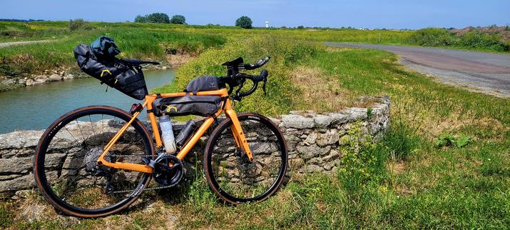 Photo d'un vélo orange posé contre un muret de pierres dans un marais. Le vélo possède des sacoches accrochées au cadre et à la selle. À droite de la photo une petite route qui jouxte le marais. Derrière le vélo, un bassin rempli d'eau.