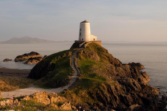 A white painted lighthouse stands on top of a rocky mound. A white path leads up to the lighthouse. The sea around the lighthouse is calm and flat. Across the sea a mountain can be seen slightly obscured by haze. The sun is setting to the right of the photo casting a golden hue across the scene.
