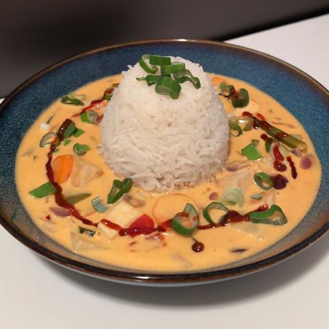 Picture of a red coconut curry with tofu, carrots, red onions, green beans and green bell pepper. In the middle of the curry, there is a cup-shaped portion of basmati rice. The meal is seasoned with spring onions and a ring of Sriracha chili sauce in a ring aroind the rice. It is served on a blue plate. The background is a neutral white table.