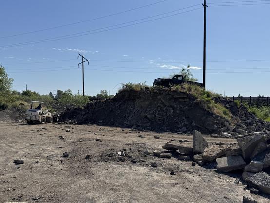 Construction site with a loaded vehicle moving debris. A pile of rubble is visible in the background, along with power lines and clear blue sky. A black pickup is parked on top of the pile