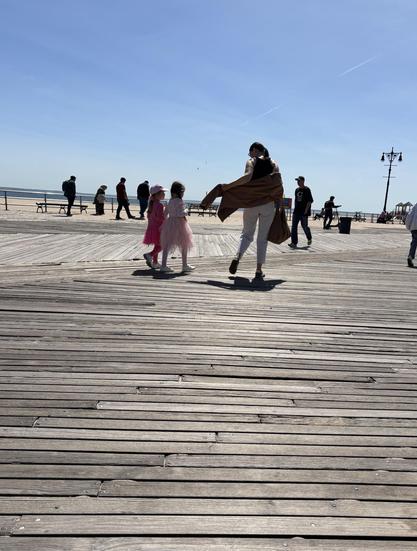 Very young sisters, each in pink tutus, follow their mother as she puts on her light jacket. They walk away from me on the boardwalk, beach and ocean in background.