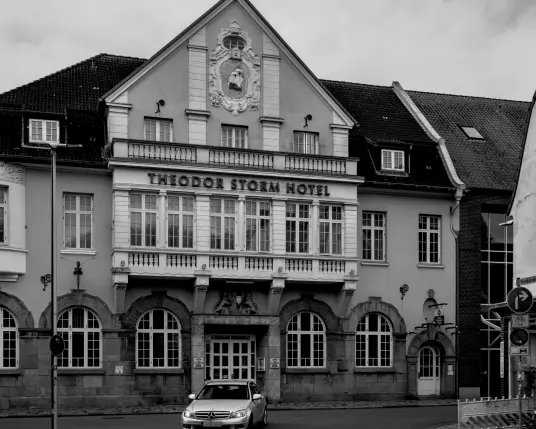 Irgendwo mitten in Husum steht das Theodor Storm Hotel, ein alter Bau mit geschwungenen Fenstern im Erdgeschoss, mit angedeuteten Säulen. Oben im Giebel ist ein großes Wappen zu sehen, reich verziert. Im Wappen segelt ein Schiff über das Meer. 
Vor dem Hotel biegt ein Auto ab in eine Seitenstraße. 
. 
Somewhere in the centre of Husum there is the Theodor Storm Hotel, an old building with curved windows on the ground floor and implied columns. At the top of the gable is a large, richly decorated coat of arms. In the coat of arms, a ship sails across the sea. 
A car turns into a side street in front of the hotel. 
.