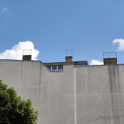 Eine breite, graue, fast fensterlose Seitenwand eines mehrstöckigen Hauses. Lediglich an der Oberkante befinden sich als Teil eines Aufbaus auf dem Dach drei kleine Fenster. Eins davon dreieckig, die anderen beiden viereckig. Von links ragt eine grüne Baumkrone ins Bild. Über drei Schornsteinen auf dem Dach des Hauses ist ein blauer Himmel mit ein paar weißen Wolken zu sehen.