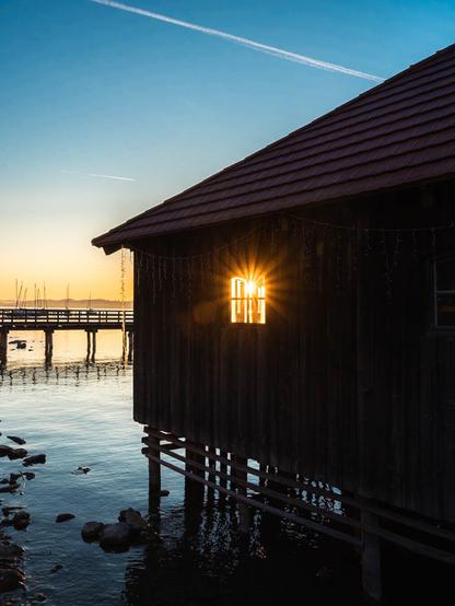 This image shows a peaceful lakeside scene at sunset. In the foreground is a wooden boathouse or cabin built on stilts over water. The structure has dark wooden walls and a brown shingled roof. A window on the side of the building is capturing the setting sun, creating a dramatic starburst effect as the sunlight shines directly through the glass.

The boathouse stands in calm water where small rocks are visible beneath the surface. To the left, you can see part of a wooden pier or dock extending into the water. In the background, there are what appear to be sailboat masts visible near the shoreline.

The sky is a beautiful gradient, transitioning from deep blue at the top to golden yellow near the horizon where the sun is setting. A single contrail from an airplane cuts across the blue sky. The overall atmosphere is serene and golden, capturing that magical moment when day transitions to evening over a tranquil body of water.