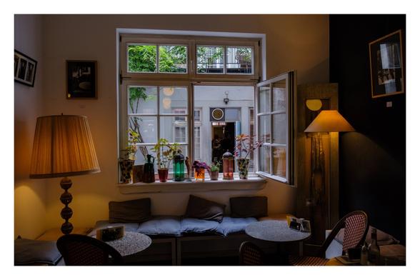 Foto im Querformat. Blich durch ein Café auf ein offenes Fenster. Im Café sind Tische, Bänke und Stühle. Links und rechts sind Stehlampen, die den Raum in warmes Licht tauchen. An der Wand hängen Bilder. Auf der Fensterbank stehen verschiedene Vasen und Flaschen in unterschiedlichen Farben. Hinter dem Fenster ist ein Hinterhof.