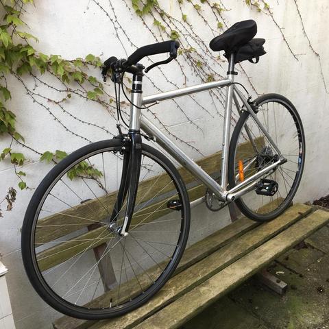 Front 3/4 picture of a hybrid bicycle, silver frame and black fork, headset, and wheels; the brake levers are mounted on the bar-ends for a funky look.