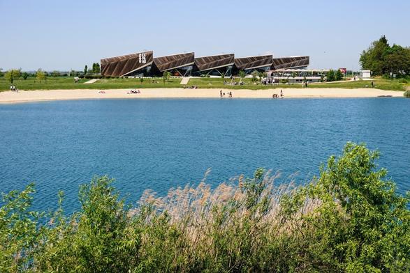 Lake viewed from slightly above, in front there are some bushes, and on the other side of the lake there is a wood-and-glass pavilion.
