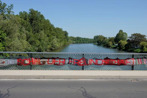 A view of a river from the middle of a bridge. Across the fence a scarf extends horizontally with writing "Schal der Freundschaft" (Scarf of Friendship).