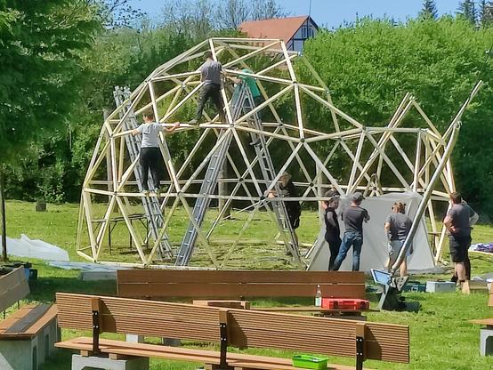 Das fast fertige Gerippe eines Geodome, einer runden Kuppel. Menschen, die beim Zusammenbau helfen. Im Vordergrund Sitzbänke, im Hintergrund das Dach des Dobelmühle Gebäudes.