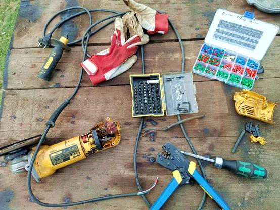 A well used outdoor workbench. There's a yellow angle grinder with the cover off, the cable removed and a piece cut off. The exposed wires have new bootlace ferrules crimped on. A crimping tool and a plastic sorter full of colourfull ferrules is nearby. There's also some gloves, a ratcheting bit screwdriver and a box of bits whose open lid holds the screws from the angle grinder.