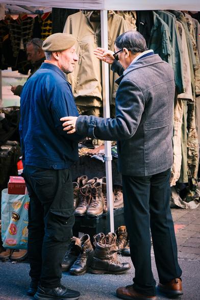 Two men standing, facing each other at an angle, in Brick Lane Market in front of a stall selling vintage clothes and boots. The man on the right is holding the right arm of the other man with his left hand and his right hand is held up in front of the other man's face with four fingers pointing at the other man's head.