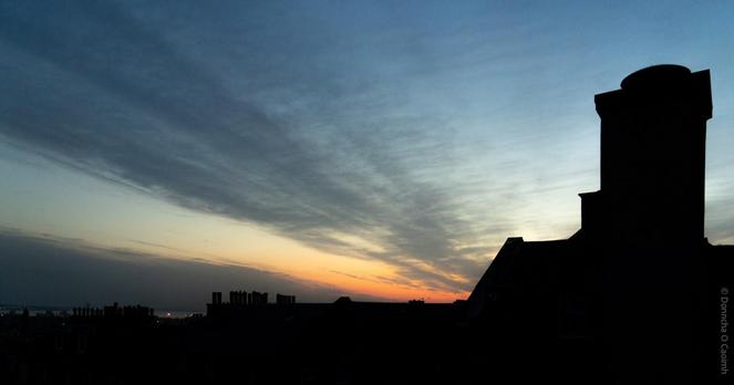 A silhouette of Edinburgh rooftops and chimneys at sunrise, with a dramatic sky streaked with clouds in blue, grey, and orange hues.