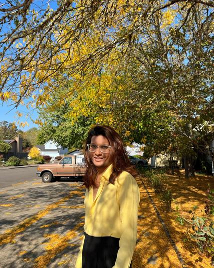 Photo of Tanveer wearing a yellow shirt and surrounded by autumn-looking trees.