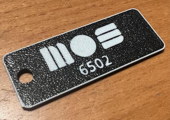 A black and white keytag with the inscription "MOS 6502." The surface appears textured, and there is a hole for attachment. The tag is placed on a wooden surface.