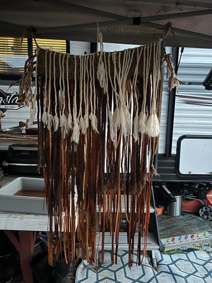Medium shot of Indigenous weaving piece hanging taking up the center of the frame. The top of the hanging is suspended by twine from a gray metal pole, likely part of a tent frame or awning structure. The hanging comprises a thick, woven band, from which hang alternating strands of thick, white rope with fluffy tassels and narrower strips of tanned leather and knotted brown cord. The visual texture is varied due to the different materials. The backdrop is a cluttered outdoor scene, including a partial view of a large RV to the right, and a workbench with various camping supplies, tools beneath and to the left. The lighting is natural daylight, with some areas in shadow.