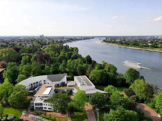 Ehemaliger Kanzlerbungalow um geben von vielen alten Bäumen. Rechts davon fährt auf dem Rhein ein Ausflugsdampfer. Bis zum Horizont Bonn.