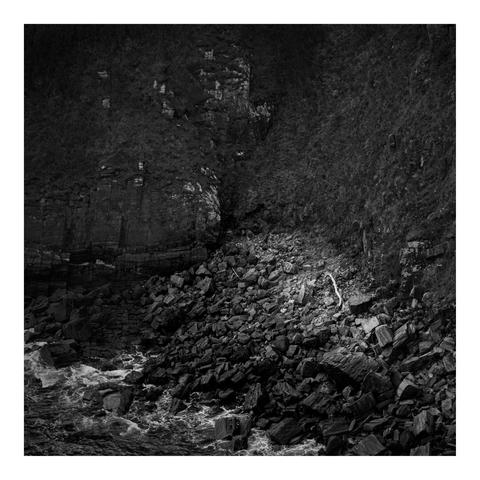 A moody black and white image captures a rugged coastline with jagged rocks, steep cliffs, and crashing waves. A pale driftwood log lies among the dark stones, contrasting sharply. The textured cliff and hillside add depth, while the turbulent sea emphasizes nature’s raw, untamed power and atmospheric isolation.