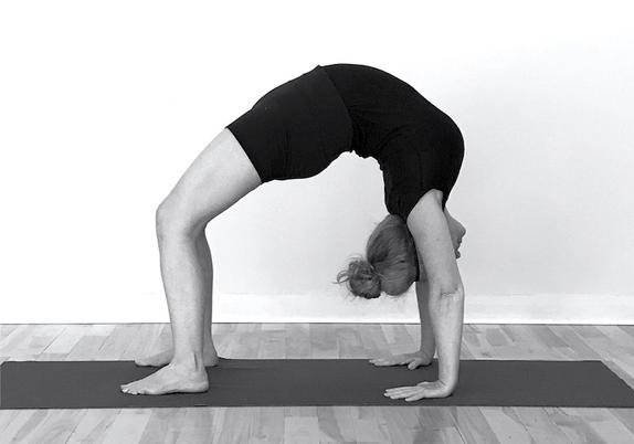 Une photo en noir et blanc montrant une yogini pratiquant la posture de la roue : jambes et bras tendues, le dos cambrée, en flexion arrière, la tête perpendiculaire au tapis