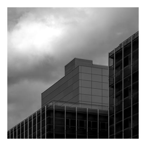 A monochrome photograph of modern buildings under a dramatic, cloud-filled sky. The architecture features clean lines, glass panels, and geometric shapes, with one central structure highlighted. The lighting emphasizes contrast between sleek surfaces and moody weather, creating a stark, minimalist urban atmosphere that evokes a sense of stillness and isolation.