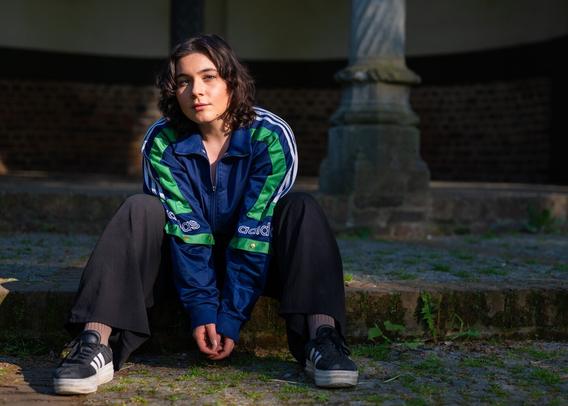 Eine junge Frau sitzt auf Stufen, sie schaut direkt in die Kamera. Sie trägt Speaker eine schwarze Hose sowie eine blaue Jacke mit etwas grün an den Ärmeln. 
Ihr Gesicht wird von der Sonne beleuchtet, ein kleiner Teil liegt im Schatten.