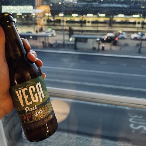 A hand holds a bottle of Vega Post IPA, a beer brewed specially for the Clarion Hotel Post in Göteborg. Through the hotel room window, the illuminated Göteborg Central Station and trains are visible in the evening light, with taxis and commuters bustling outside. The scene captures a relaxing moment enjoying a local craft beer with a view of the city’s main transport hub.