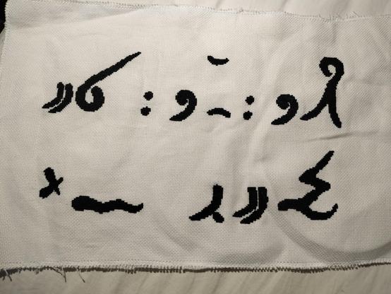 a cross stitch in black floss on a white hardanger weave. it has two rows of symbols, which mostly consist of various dots, swirls and curved lines.