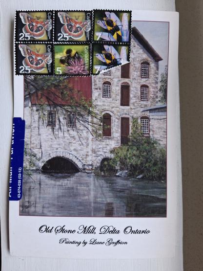 A postcard with a painting of a large stone mill set in water. The mill looks about 3 stories high with grey stone and red coloured wooden roof arches. At the top of the postcard are postage stamps with a brightly red coloured moth, a bumblebee, and a yellow and black beetle.