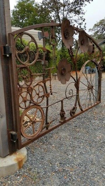 A gate made of steel junk. There's a few disc harrows, a large spoked steel rim wheel, various cogs and pulleys, an auger, an axewith a curve dhandle and lots more welded into an outer profile frame.