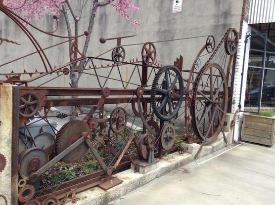 A different steel junk gate or perhaps just part of a fence. This one is mostly gearwheels, some chains and various links.There's a frame, but the add-ons exceed the frame.