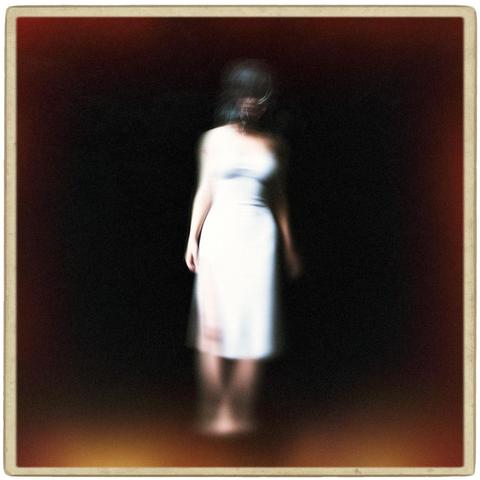 Full-length portrait of a woman with dark hair and wearing a white dress. The woman is blurred considerably from motion. The background is completely black. The image is square and surrounded by a grungy photo border. The edges of the image are fogged orange and red from light leaks.