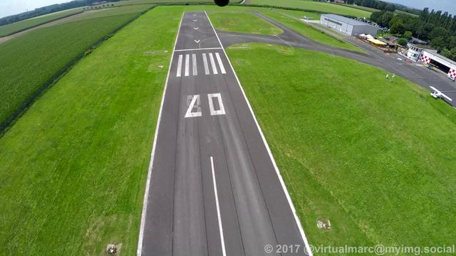 A photo of a runway for planes from below a gyrocopter down to the ground. There is green grass around the runway, part of some hangers and the tower can be seen on the right.