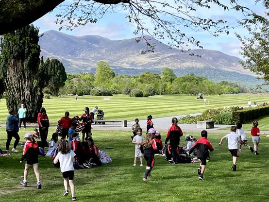 Foreground shows several children running in different directions. A sweeping branch with leaves just budding out caps the frame. Small mountains in the background.