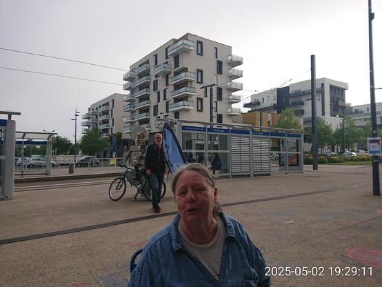 Femme en t-shirt et veste jeans parlant à la terrasse du Café des Images, devant le drapeau de l'asso Hérouelibre, un homme descenu de son vélo. Au second pan, la station de tramway et quelques immeubles alors que la brume descend sur la ville.