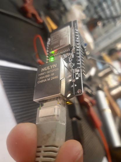Two fingers hold an ethernet cable plugged into a small electronics board with dense circuitry and a number of I/O pins.