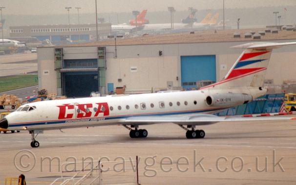 Side view of a white, twin engined jet airliner with the engines mounted on the sides of the rear fuselage, taxiing from right to left.
The plane is mostly white, with large red "CSA" titles on the sides of the forward fuselage, with white "Czechslovak Airlines" text just below on a thin blue stripe running along the body.
A pair of red triangles separated by a blue triangle adorn the leading edge of the tail, with red piping underneath.
The engine pods on the rear fuselage are grey, with the registration "OK-EFK" in black.
A white building with a flat, sandy brown roof and blue roller doors stretches across most of the frame from the right, with the tails of other jet airliners next to a brown building in the distance.