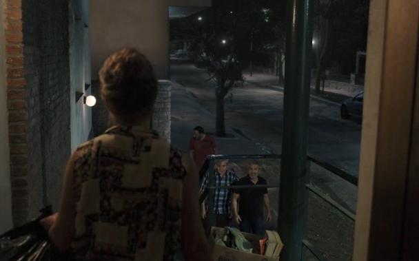 Over shoulder shot outdoor shot of a woman on top of some stairs in front of a building entrance, looking down on 3 men who are just arriving.
There are lamps in the foreground that illuminate the building and the sidewalk in a warm light. Most of the background, however, looks noticeably cooler and the asphalt of the street and sidewalk is lacking texture. It looks like a CG replacement of whatever was there when the scene was shot and the line between real plate and CG is exactly where a metal railing is located, like a matte created in compositing.