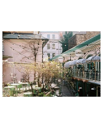 Café tables in light green, everything a little Juigendstil like. Tree in the middle, could be Magnolie, not blooming yet. Light chains crossing the picture, some sun shades, rose colored back of a building in the background, stairways going downstairs, balcony levels... from everything a little to much