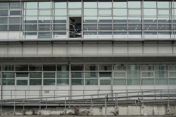 Weißgraue Fassade mit zwei Fensterreihen. In ersten Stock ist ein einziges der Fenster geöffnet. Im Vordergrund Geländer und eine Zugangsrampe.