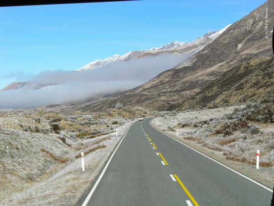 A road leads the eye into the picture. On the immediate left and right is brown grass and shrubs, lightly dusted with frost. Ahead, a mountain range descends from the right to meet the road, which curves out of sight. Past the first flank of the mountain, a dense white, very low  cloud bank obscures the view apart from some snow-capped peaks above and behind.