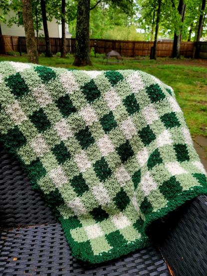 A close-up shot captures a cozy, hand-crocheted blanket draped over a dark, textured surface, likely part of outdoor furniture. The blanket features a charming gingham check pattern in three shades of green: a deep forest green forming the solid checks and the border, a lighter sage green, and a creamy off-white creating the alternating squares. The texture of the crochet is evident, with small, raised stitches forming the fabric.

The blanket is positioned diagonally across the frame, with a portion cascading down the side of the furniture, revealing its woven texture. The background is softly blurred, showcasing an outdoor setting with green grass, several trees with visible trunks and foliage, and a wooden fence in the distance. Hints of a light-colored house are visible through the trees on the left side of the frame. The overall lighting suggests it's daytime, with natural light illuminating the scene, casting soft shadows.