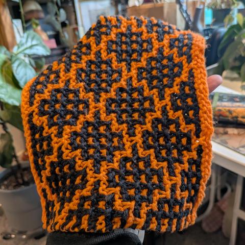 A knit orange washcloth with black geometric diamond motifs. Mosaic knitting techniques. Variety of houseplants in the background.