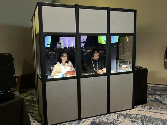 Two interpreters with headsets inside a sound-proof booth. Presentation screens are reflected in the glass.