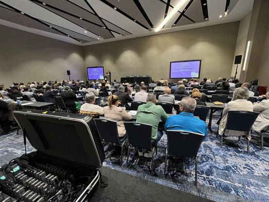 A full conference room viewed from the back. A speaker is on stage, there are two large screens showing a presentation. In the foreground there is a case with electronic equipment on a table.
