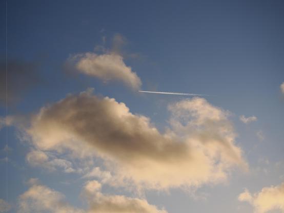The image shows a clear blue sky with some scattered clouds. There is also a white contrail, likely from an airplane, extending across the sky.